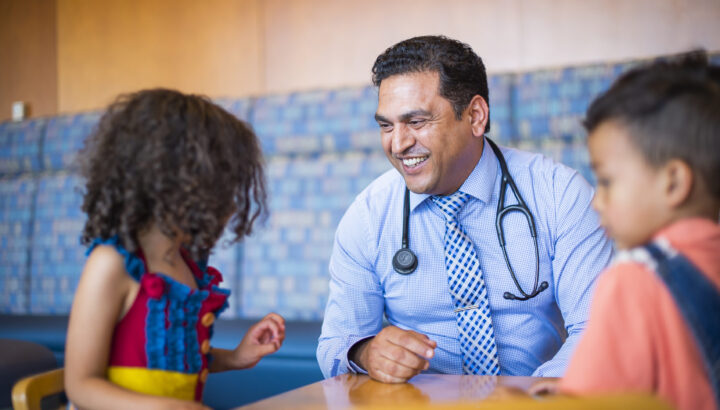 Smiling male doctor wearing a stethoscope around his neck, sitting with a little girl and boy.