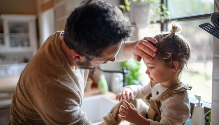 Father checking forehead of sick toddler daughter indoors in kitchen at home.