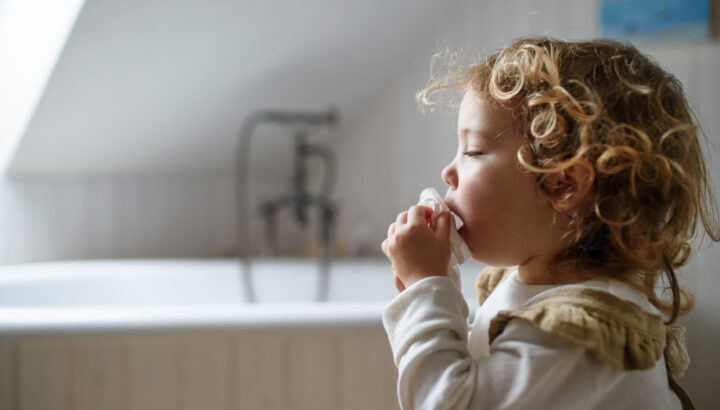 Small sick girl with cold at home sitting on ground, sneezing. Copy space.