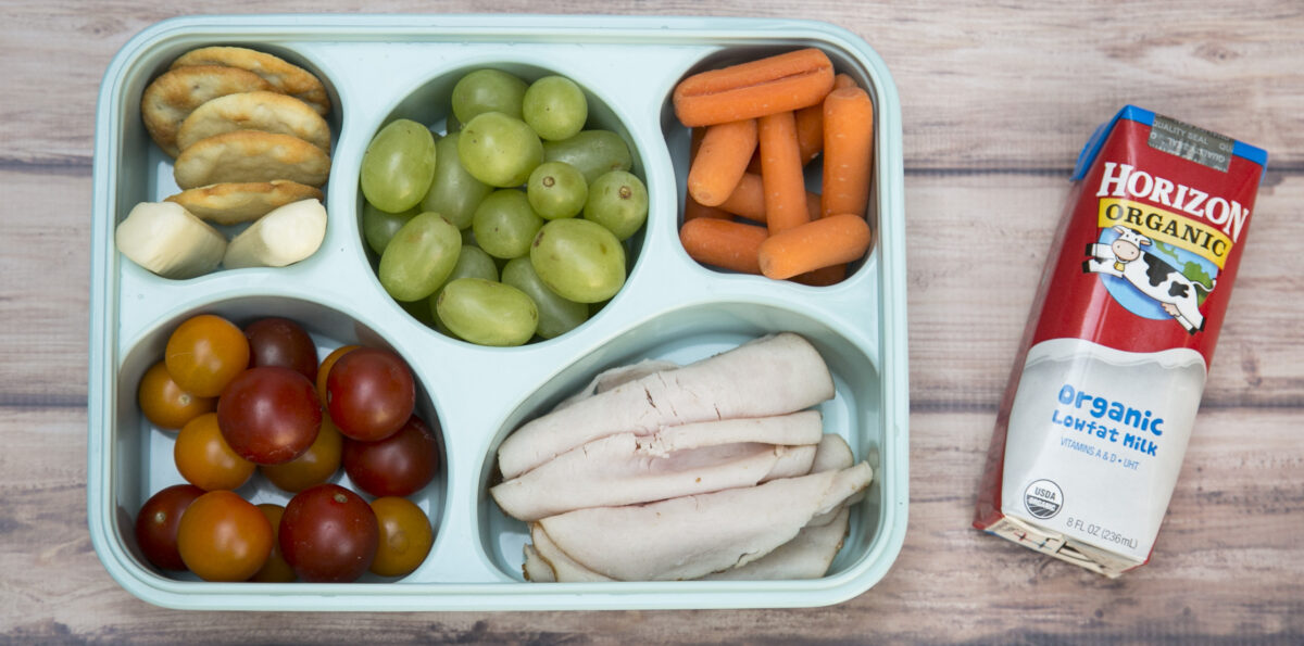 A bento box featuring rolled-up turkey slices, fruits, vegetables and a carton of milk.