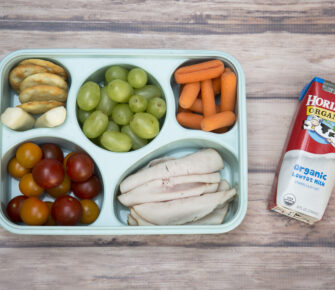 A bento box featuring rolled-up turkey slices, fruits, vegetables and a carton of milk.