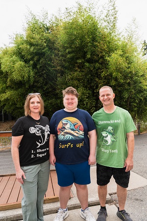 Liam, mom Bev and dad Graeme stand smiling outside the Outer Reef exhibit in Frisky Shark apparel that Liam designed.