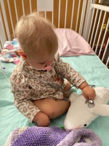 Toddler in a hospital crib with a pacifier in her mouth using a stethoscope to check the vitals on her stuffed animal whale