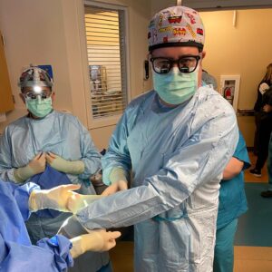 Dr. Escobar in the operating room wearing blue surgical scribs, a green mask and a surgical cap