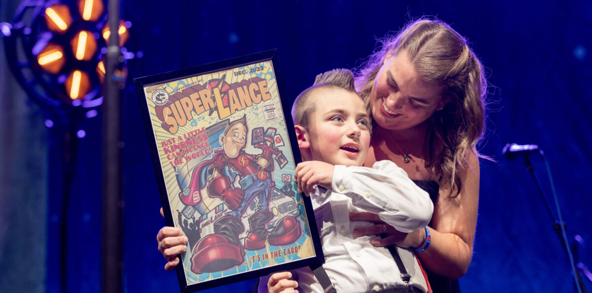 A young boy in a tuxedo and cape stands onstage holding a comic-style portrait of a superhero, and is embraced from behind by his mother.