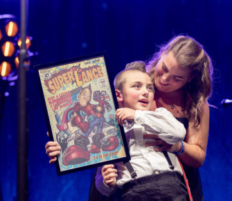 A young boy in a tuxedo and cape stands onstage holding a comic-style portrait of a superhero, and is embraced from behind by his mother.