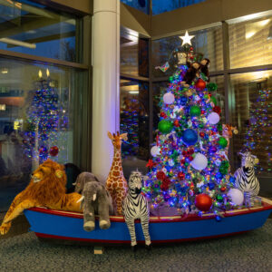 A lit Christmas tree is decorated with Mary Bridge Children’s kites and kid-crafted ornaments, surrounded by giant stuffed animals sitting in a wooden boat. 