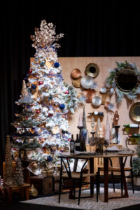 A wine country-themed Christmas tree is lit in front of a well-stocked wine cabinet and small metal dining room table.