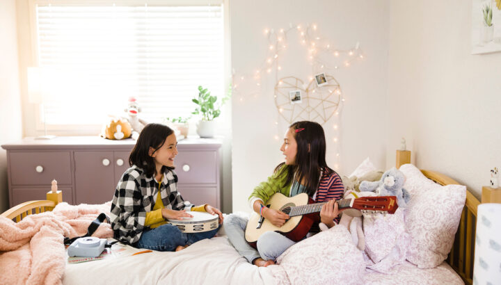 Two tween girls playing a tambourine and guitar in light, airy room while sitting on bed.