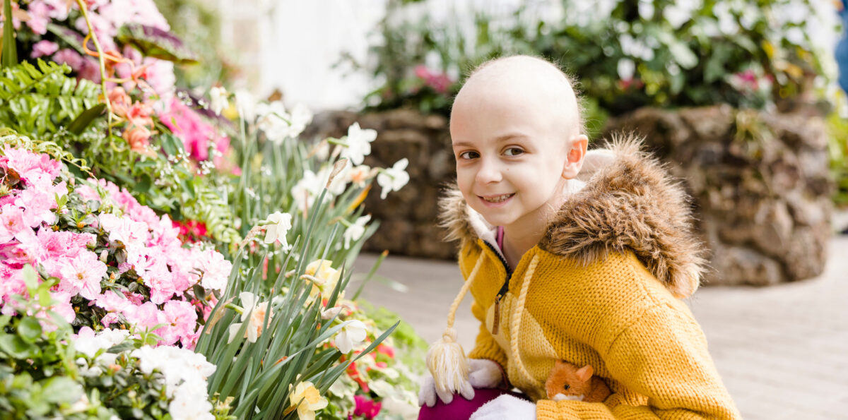 Patient Ambassador Aile smiles next to flowering bushes.