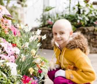 Patient Ambassador Aile smiles next to flowering bushes.