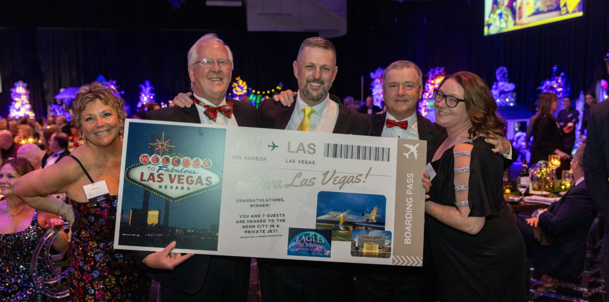 5 glamorously dressed winning bidders, surrounded by Christmas trees, hold up a giant airline ticket from Gig Harbor to Las Vegas.