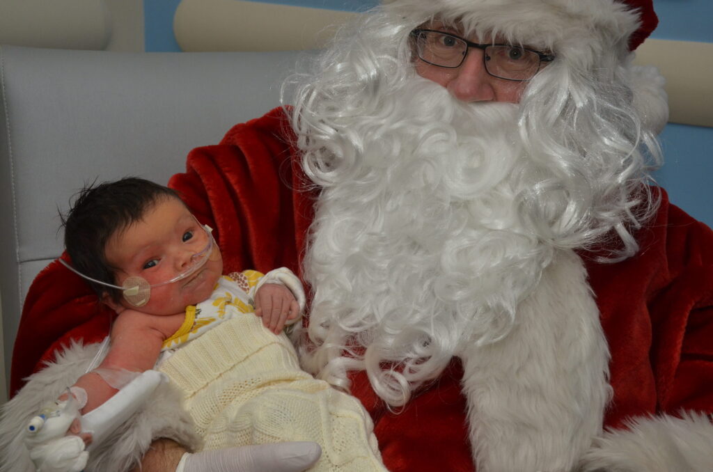 Santa wears glasses and holds a smiling brunette baby in a yellow blanket with oxygen tubes and an IV placement.