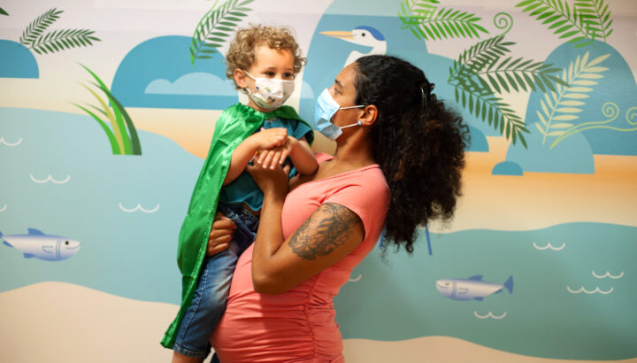 Masked mother, holds her masked son, while he wears a green cape.