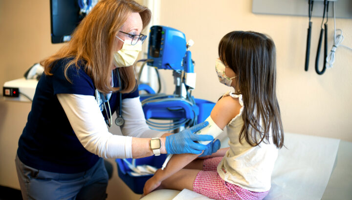 A masked nurse, tending to a masked young girl on an examination table.