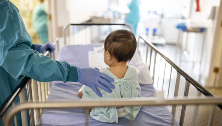 Infant sitting on a gurney, with a doctor's hand on their back.