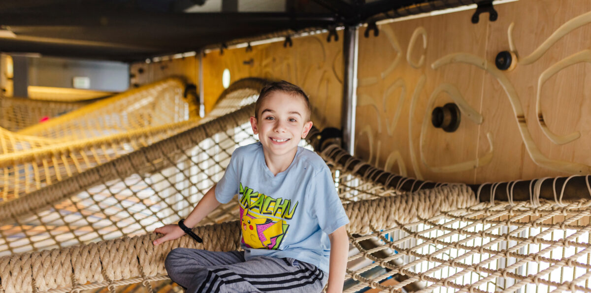 Oakley poses for a photo in a play structure, sitting on a suspended cargo net.