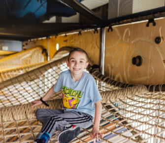 Oakley poses for a photo in a play structure, sitting on a suspended cargo net.