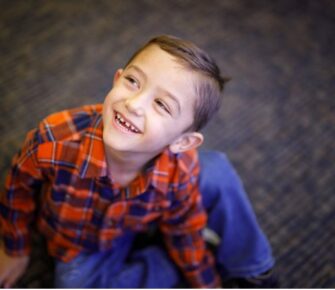 Young boy in a red-and-blue shirt sitting on the floor, looking up and smiling