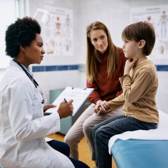 Doctor talks to child, who is touching his throat, and his mother