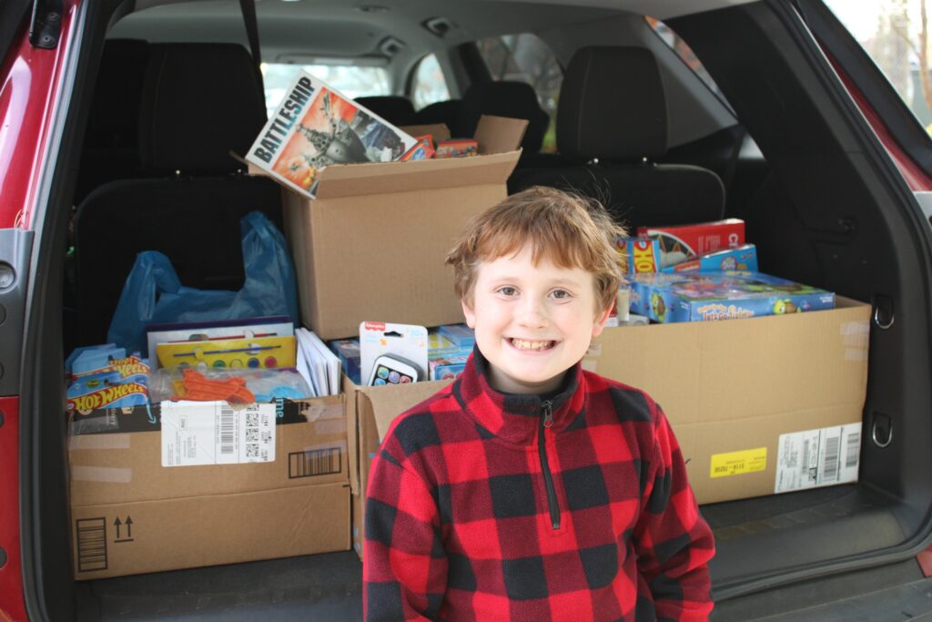 A young redhead boy in a red plaid sweater smiles in front of an open Subaru trunk, filled with cardboard boxes of donated toys.