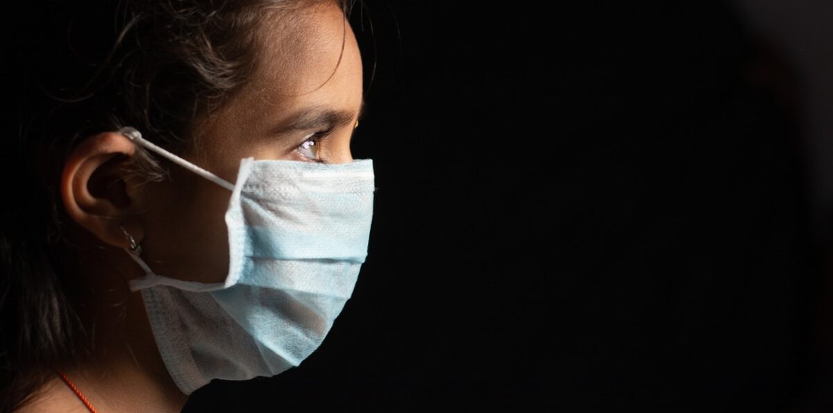 Young teenager girl with medical face mask in dark room at home quarantine due to covid 19 or coronavirus outbreak - concept of hope and fight to end virus crisis