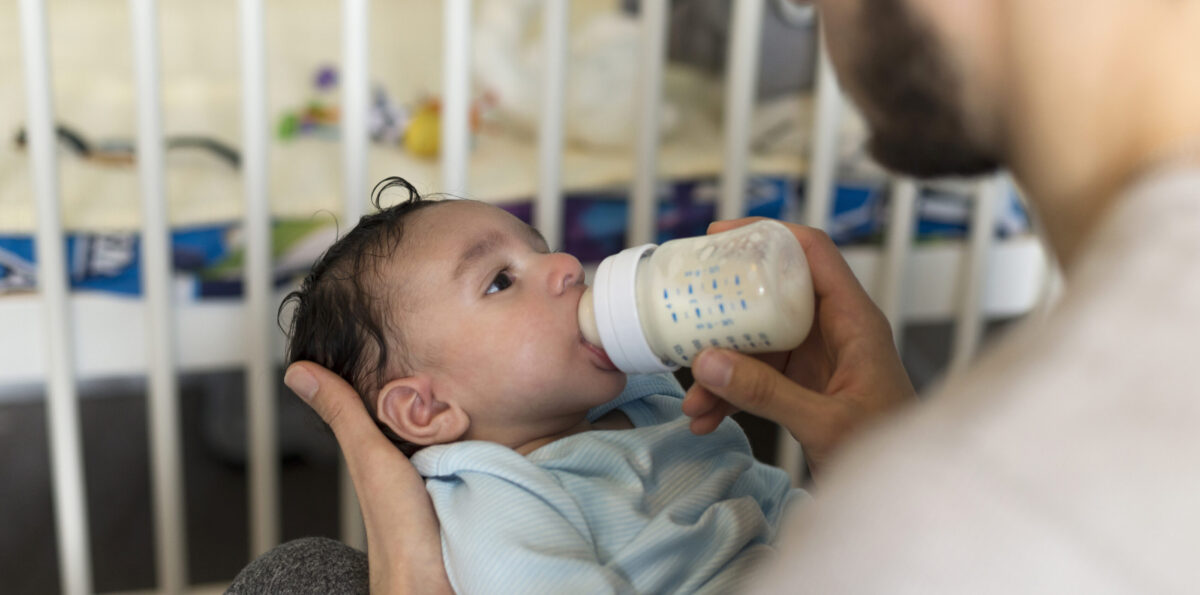 Young father is sitting with his baby son in his lap. He is feeding him a bottle of milk.