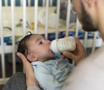 Young father is sitting with his baby son in his lap. He is feeding him a bottle of milk.