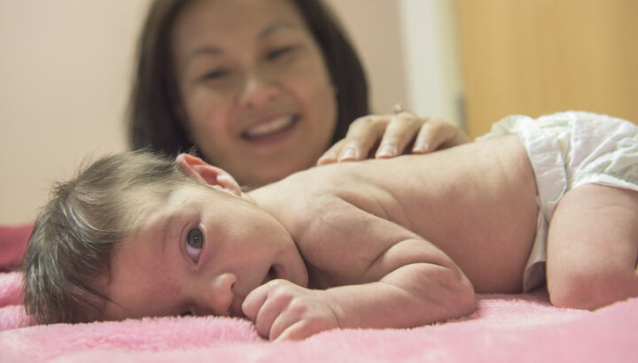 Infant, laying on their tummy on a pink blanket, with a smiling woman in the background.