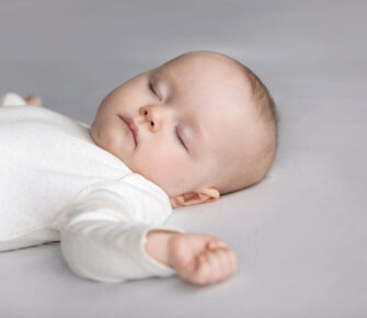 Calm silent adorable baby in white bodysuit sleeping on bed. Cute few month infant kid resting on pale sheet, linen. Child care, bedtime, daily routine concept.