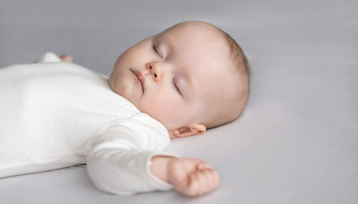 Calm silent adorable baby in white bodysuit sleeping on bed. Cute few month infant kid resting on pale sheet, linen. Child care, bedtime, daily routine concept.