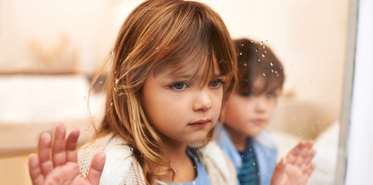 Shot of two unhappy-looking young children looking out a window on a rainy day