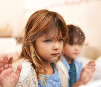 Shot of two unhappy-looking young children looking out a window on a rainy day