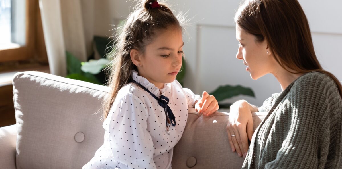 Upset small schoolgirl having trustful conversation with compassionate young mother, sitting together on sofa. Wise mommy comforting soothing little child daughter, overcoming problems at home.