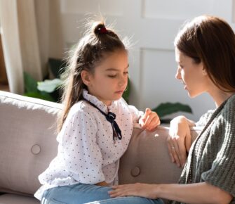 Upset small schoolgirl having trustful conversation with compassionate young mother, sitting together on sofa. Wise mommy comforting soothing little child daughter, overcoming problems at home.