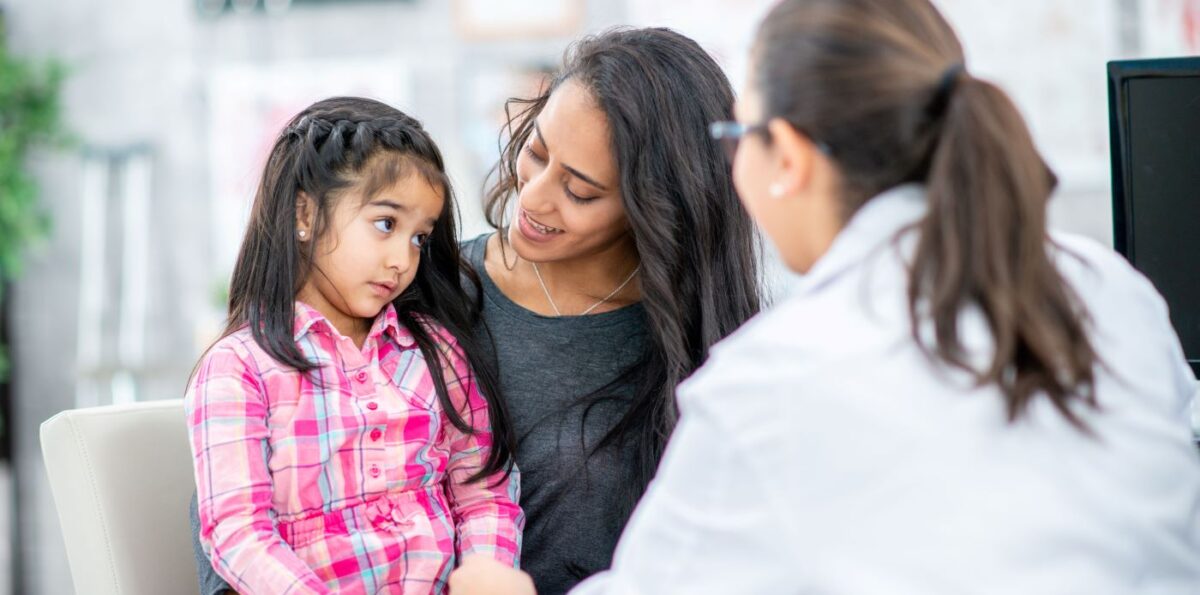 A female doctor talks to a young female patient who sits, listening, with her female caregiver by her side.