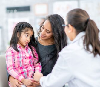 A female doctor talks to a young female patient who sits, listening, with her female caregiver by her side.