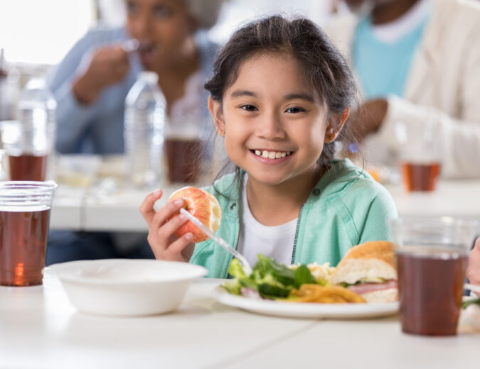 Cute young girl smiles while having a healthy meal in a cafeteria. She is eating an apple.