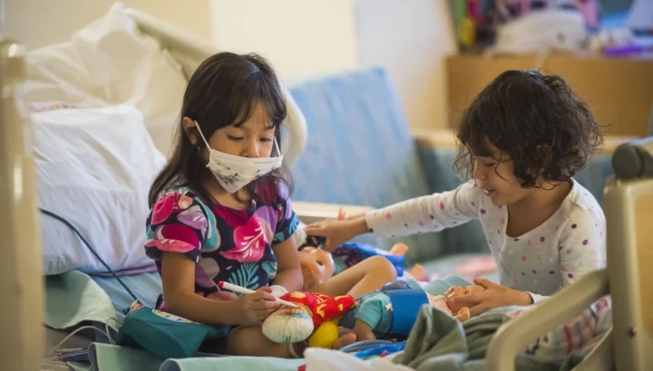 Two toddler girls, one with a mask on, sitting in a hospital bed playing.