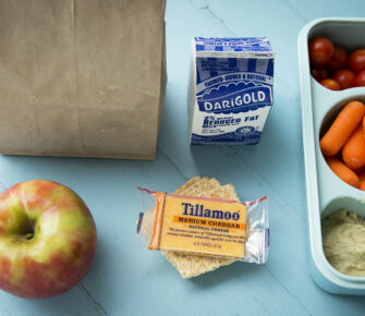 Healthy school lunch laid out showing milk, apple, cheese and crackers, humus and vegetables.