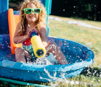 Girl in sunglasses sitting in a kiddie pool with a big squirt blaster.