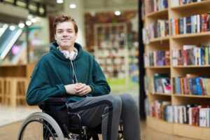 Content handsome young student with headphones on neck siting in wheelchair and looking at camera in modern library or bookstore