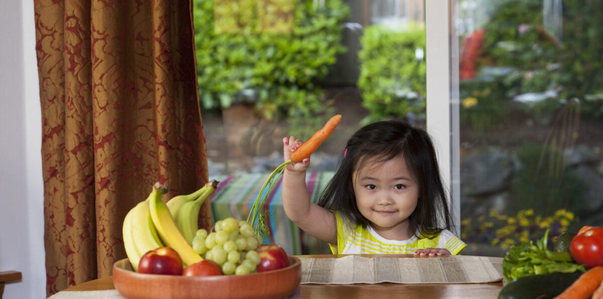 Little girl holding a carrot next to a bowl of fresh fruits.