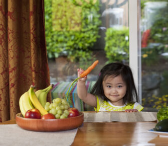 Little girl holding a carrot next to a bowl of fresh fruits.