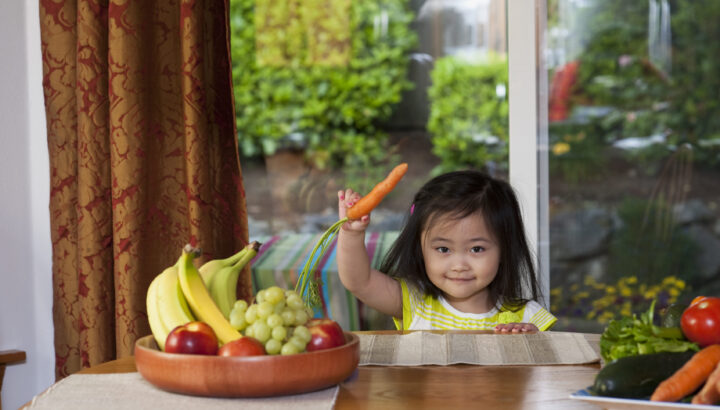 Little girl holding a carrot next to a bowl of fresh fruits.