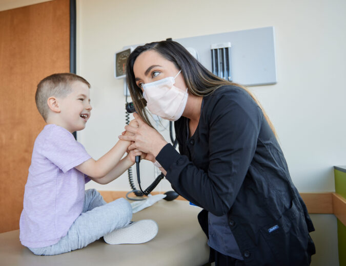 Young boy sitting on exam room table, smiling and looking into health care provider's ear with an otoscope.