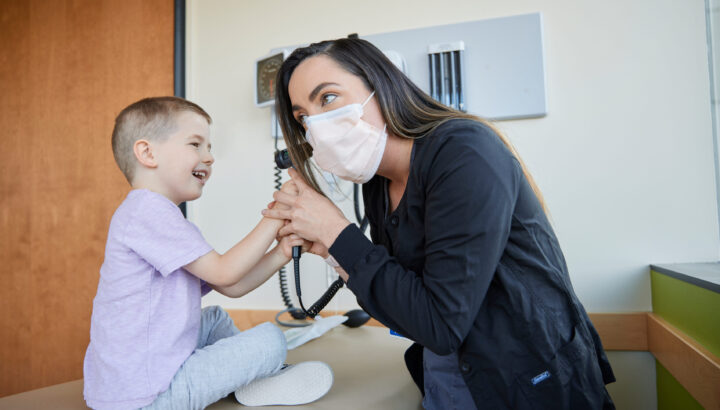 Young boy sitting on exam room table, smiling and looking into health care provider's ear with an otoscope.