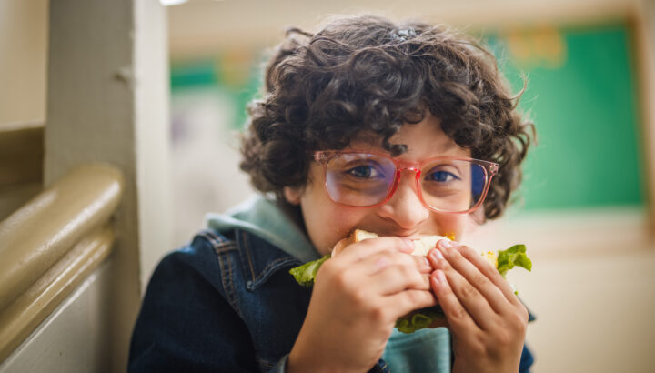 Young boy wearing jean jacket and glasses, sitting on a staircase at school biting into a sandwich.