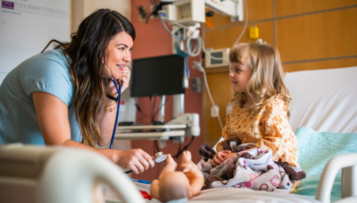 Woman in hospital room with little girl sitting on bed, pretending to use stethoscope to check on play doll.