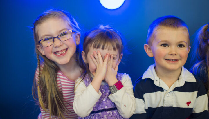 Three smiling kids sitting in the Mary Bridge Children's playroom. The youngest girl sits on an older girl's lap with her hands covering her face.